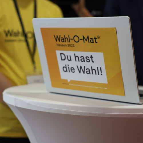 Auf einem Stehstich mit weißer Husse steht ein Laptop. Auf diesem klebt ein gelbes Schild mit dem Aufdruck "Wahl-O-Mat" Hessen 2023. Du hast die Wahl!