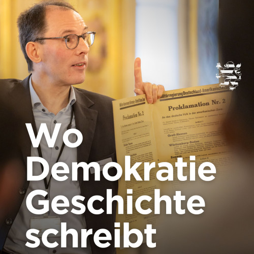 Ein Gästeführer des Landtages steht im historischen Musiksaal und hält eine Kopie der Proklamation in die Höhe. Auf dem Bild ist der Schriftzug "Wo Demokratie Geschichte schreibt. 80 Jahre Hessen" zu lesen.