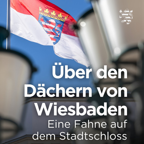 Die Fahne auf dem Dach des Wiesbadener Stadtschlosses ist im Bild zu sehen. Unscharf im Vordergrund erkennbar sind Straßenlaternen, die sich auf dem Platz vor dem historischen Gebäude befinden. Auf dem Bild ist der Schriftzug "Über den Dächern von Wiesbaden. Eine Fahne auf dem Stadtschloss" zu lesen.