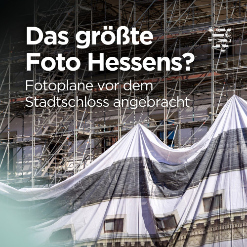 Die neue Fotoplane am Stadtschloss ist im Bild zu sehen. Entstanden ist das Bild während der Anbringung der Plane: Das Baugerüst darunter ist zu erkennen und die Plane hängt noch in Falten in die Tiefe. Der Schriftzug "Das größte Foto Hessens? Fotoplane vor dem Stadtschloss angebracht" ist zu lesen.