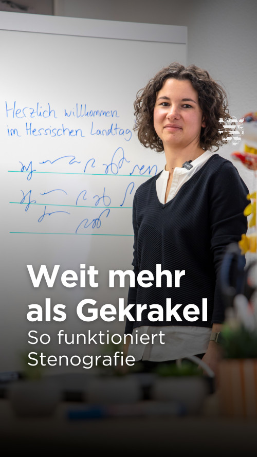 Parlamentsstenografin Rebecca Recebs ist im Bild zu sehen. Sie steht vor einem Flipchart in ihrem Büro, auf dem der Schriftzug "Herzlich willkommen im Hessischen Landtag" zu lesen ist. Den Satz hat sie außerdem in Stenografie übersetzt.