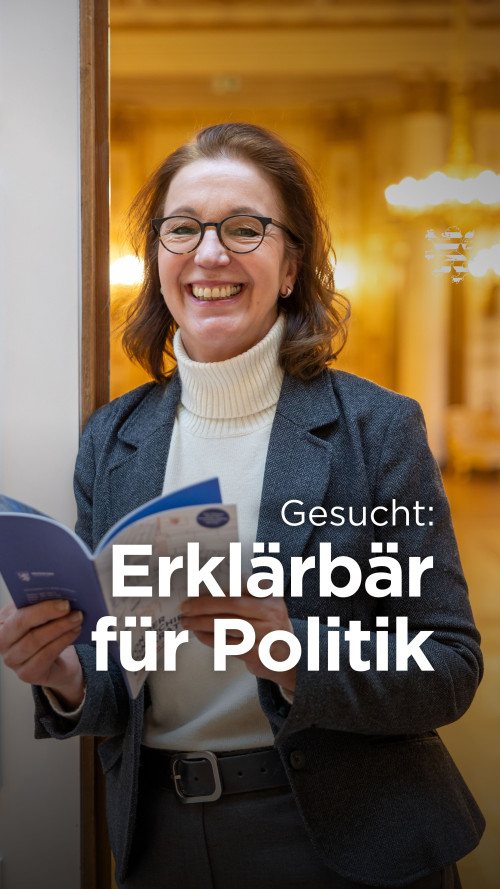 Die stellvertretende Leiterin für Politische Bildung ist im Bild zu sehen. Sie hält eine Broschüre in den Händen und lehnt an einer Tür. Unscharf im Hintergrund zu sehen ist der Musiksaal des Landtages. Der Schriftzug "Gesucht: Erklärbär für Politik" ist zu lesen.