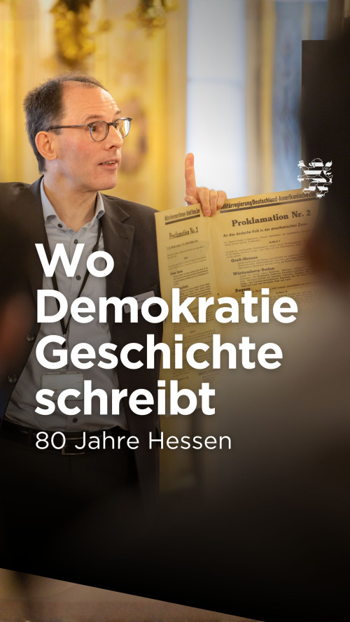 Ein Gästeführer des Landtages steht im historischen Musiksaal und hält eine Kopie der Proklamation in die Höhe. Auf dem Bild ist der Schriftzug "Wo Demokratie Geschichte schreibt. 80 Jahre Hessen" zu lesen.