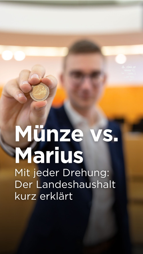 Ein Mann ist unscharf im Bild zu sehen. Er trägt ein Sakko und eine Brille und steht im Plenarsaal des Landtages, dessen typische hölzerne Rückwand erkennbar ist. Er hält eine Zwei-Euro-Münze in Richtung Kamera. Die Münze ist scharf.