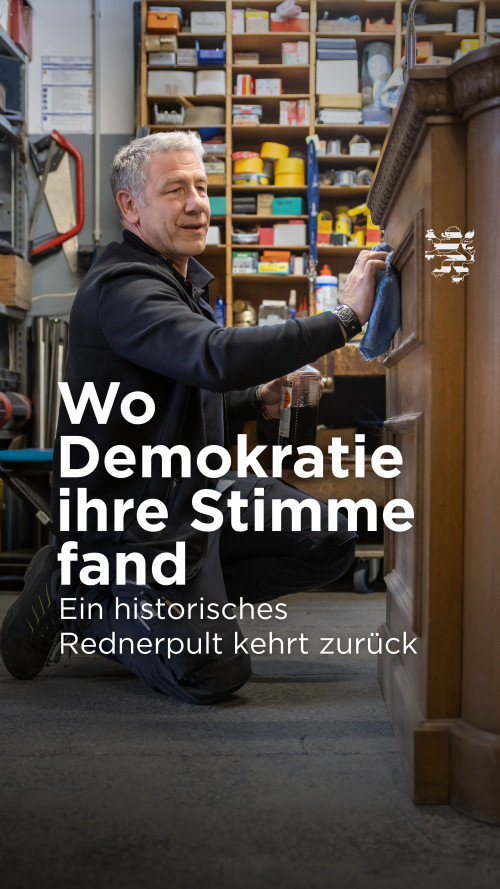 Das Bild zeigt einen Haustechniker des Landtags in einer Werkstatt. Er hält einen Lappen in der Hand und reibt damit über das Holz des historischen Rednerpults. Der Schriftzug "Wo Demokratie ihre Stimme fand. Ein historisches Rednerpult kehrt zurück" ist zu lesen.