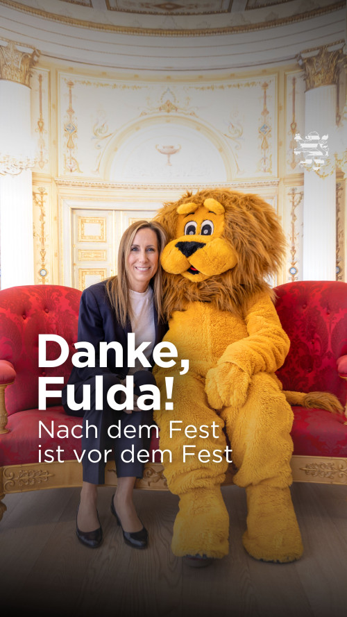 Landtagspräsidentin Astrid Wallmann sitzt zusammen mit Landtagsmaskottchen Leo auf dem Plaudersofa. Im Hintergrund ist der Musiksaal im Stadtschloss erkennbar. Der Schriftzug "Danke, Fulda! Nach dem Fest ist vor dem Fest" ist zu lesen.