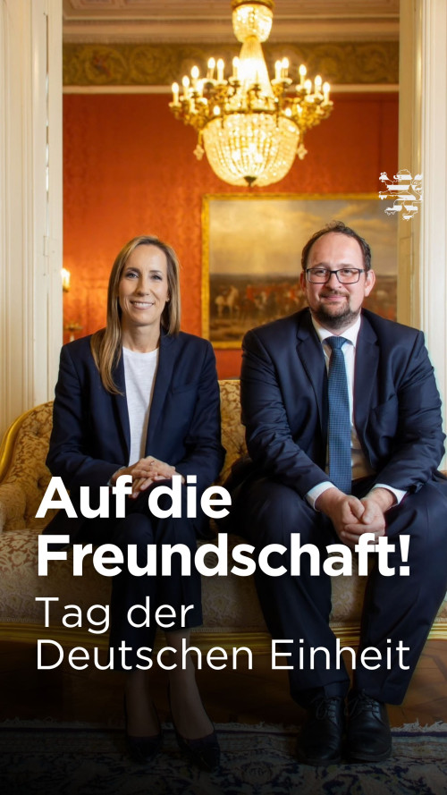 Landtagspräsidentin Astrid Wallmann und der Thüringer Landtagspräsident Thadäus König sitzen auf einem Sofa. Im Hintergrund ist der historische Präsidentensalon im Stadtschloss zu erkennen. Der folgende Schriftzug ist zu lesen: "Auf die Freundschaft! Tag der Deutschen Einheit".