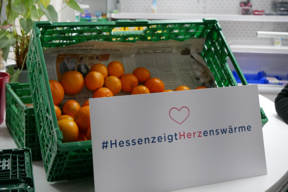 Ein Schild mit #HessenzeigtHerzenswärme vor einer Kiste mit Obst