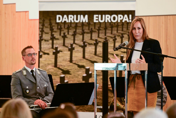 Astrid Wallmann steht an einem Podium und spricht ins Mikrofon. Im Hintergrund sitzt ein Musiker des Heeresmusikkorps Kassel.