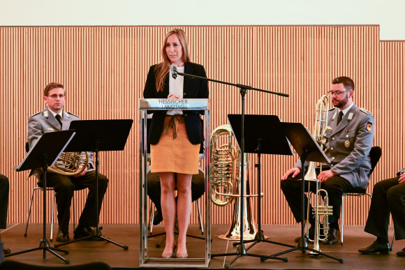 Astrid Wallmann steht an einem Podium und spricht ins Mikrofon. Im Hintergrund sitzen Musiker des Heeresmusikkorps Kassel.