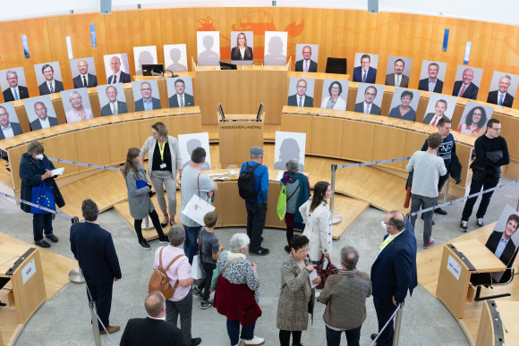 Besucherinnen und Besucher im Landtag. Auf den Sitzen sind Bilder der jeweiligen Abgeordneten aufgestellt.