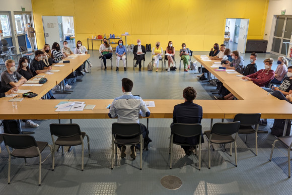 Schülerinnen und Schüler sowie Abgeordnete sitzen an Tischen, die in Hufeisenform angeordnet sind.