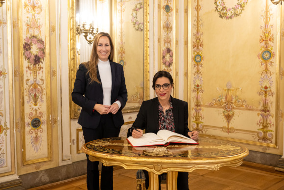 Landtagspräsidentin Astrid Wallmann steht neben der Außenministerin, die an einem Tisch sitzt und in das Gästebuch schreibt.