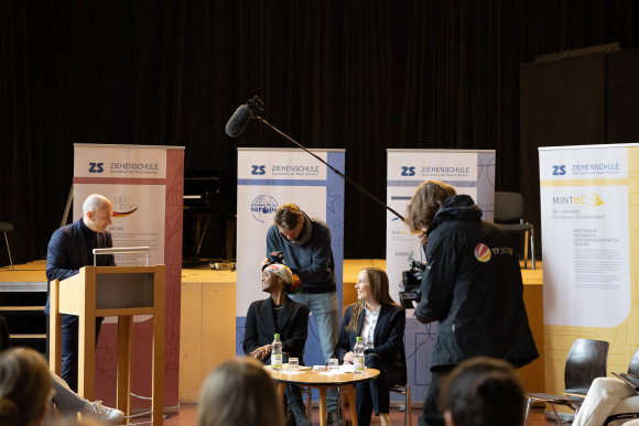Landtagspräsidentin Astrid Wallmann und die Preisträgerin des Hessischen Friedenspreises Ilwad Elman sitzen an einem kleinen Tisch in der Aula. Ein Redner am Pult spricht zu ihnen.
