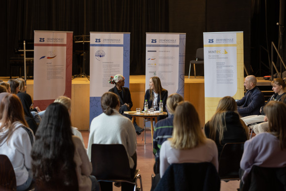Schülerinnen und Schüler sitzen im Vordergrund. Landtagspräsidentin Astrid Wallmann und die Preisträgerin des Hessischen Friedenspreises Ilwad Elman sitzen an einem kleinen Tisch in der Aula der Ziehenschule.