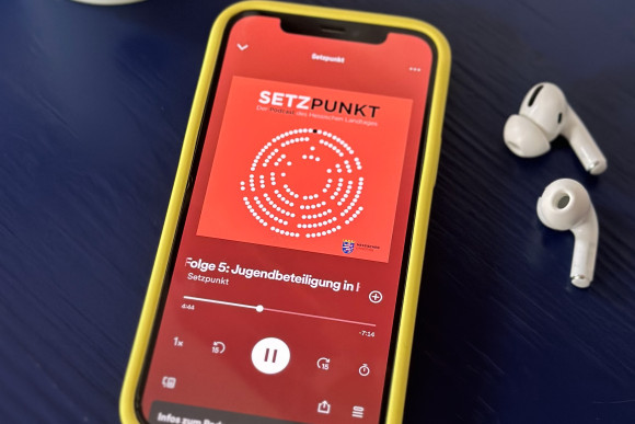 Auf einem dunkelblauen Holzuntergrund liegt ein Smartphone. Auf dem Bildschirm ist die Anzeige des laufenden Podcasts „SETZPUNKT“ zu sehen. Neben dem Smartphone liegt ein paar weiße Kopfhörer, oben links in der Ecke des Bildes steht eine weiße Kaffeetasse mit dem Wappen des Landtages, im Anschnitt zu sehen.
