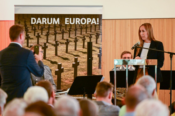 Astrid Wallmann steht an einem Podium und spricht ins Mikrofon. Im Hintergrund sitzen Musiker des Heeresmusikkorps Kassel. Vor der Bühne macht jemand ein Foto mit seinem Handy.