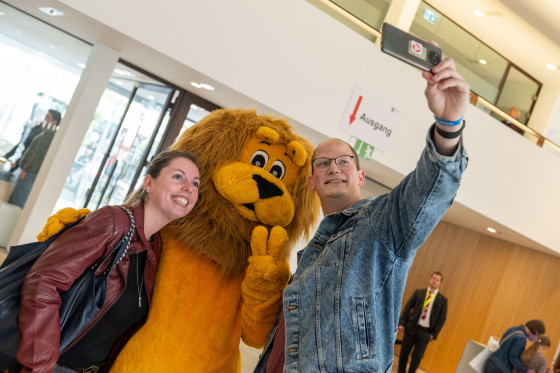 Eine Besucherin und ein Besucher machen ein Selfie mit dem Löwen-Maskottchen