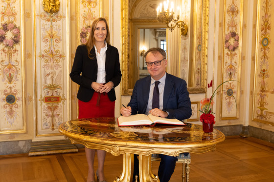 Astrid Wallmann steht neben dem Botschafter der Tschechischen Republik. Dieser sitzt an einem Tisch und schreibt in das Gästebuch.