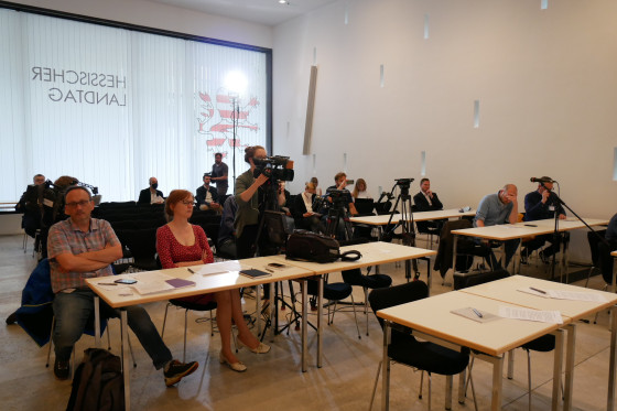 Journalistinnen und Journalisten sitzen an Tischen und hören zu. Eine Journalistin bedient eine Kamera. Weitere Mikrofone und Kameras sind im Raum verteilt.