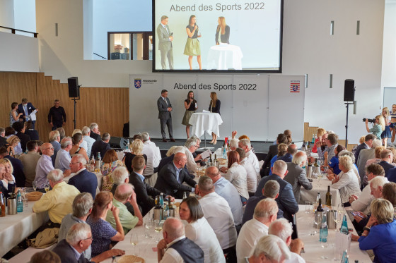 Im Hintergrund stehen Boris Rhein, Astrid Wallmann und eine weitere Person auf einer Bühne. Im Vordergrund sitzt das Publikum an Tischreihen.