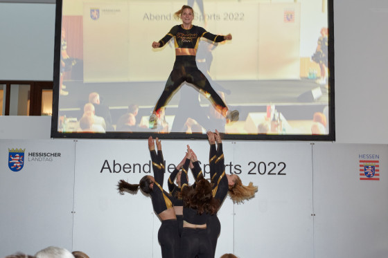 Cheerleaderinnen werfen als Teil einer Choreographie eine Cheerleaderin in die Luft. Im Hintergrund ist ein Monitor.