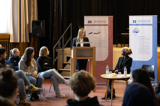 Landtagspräsidentin Astrid Wallmann spricht am Podium in der Aula der Ziehenschule. Die Preisträgerin des Hessischen Friedenspreises Ilwad Elman sitzt an einem kleinen Tisch daneben.