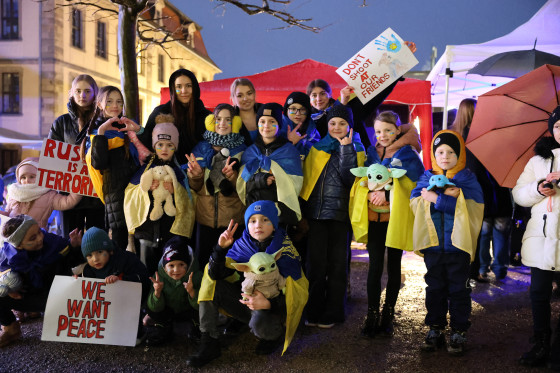 Kinder und Erwachsene stehen zusammen und tragen ukrainische Flaggen um ihre Schultern. Sie halten außerdem Plakate mit Botschaften hoch.