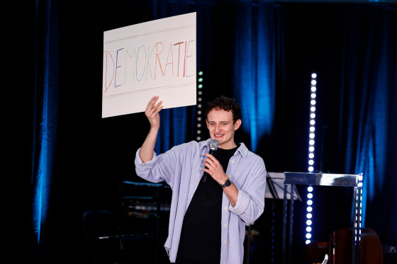 Checker Julian hält Schild "Demokratie" hoch