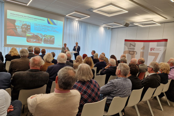 Zwei Personen vor Leinwand mit Zuschauern auf Stühlen