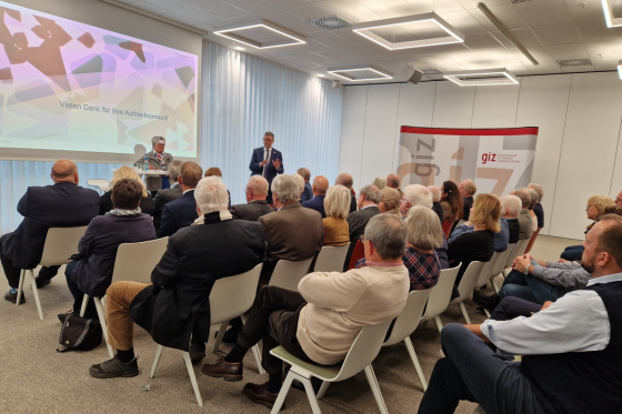 Zwei Personen vor Leinwand mit Zuschauern auf Stühlen